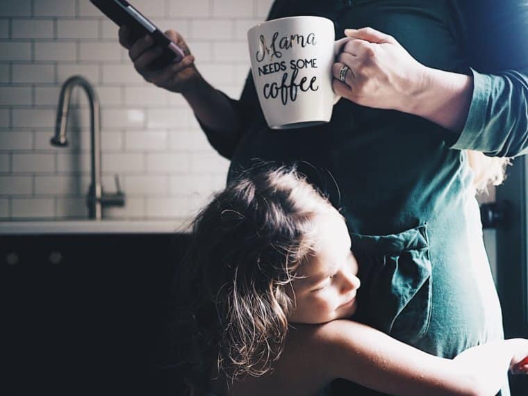 Mom looking at phone and holding coffee while little girl hugs her in post about handling stress in motherhood